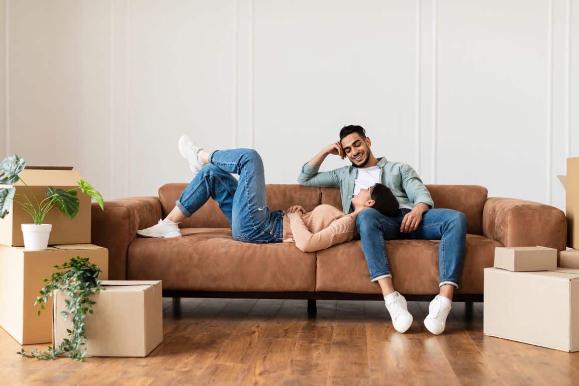 Couple relaxing on leather couch in new home surrounded by cardboard boxes.